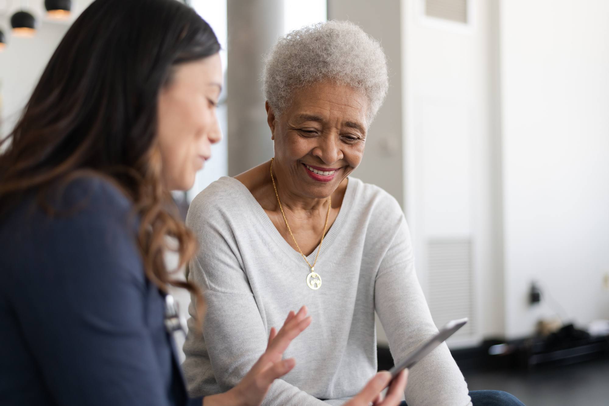 A young doctor and an elderly patient are indoors at the woman's home. The doctor is talking to the woman while holding a tablet computer. They're both smiling.